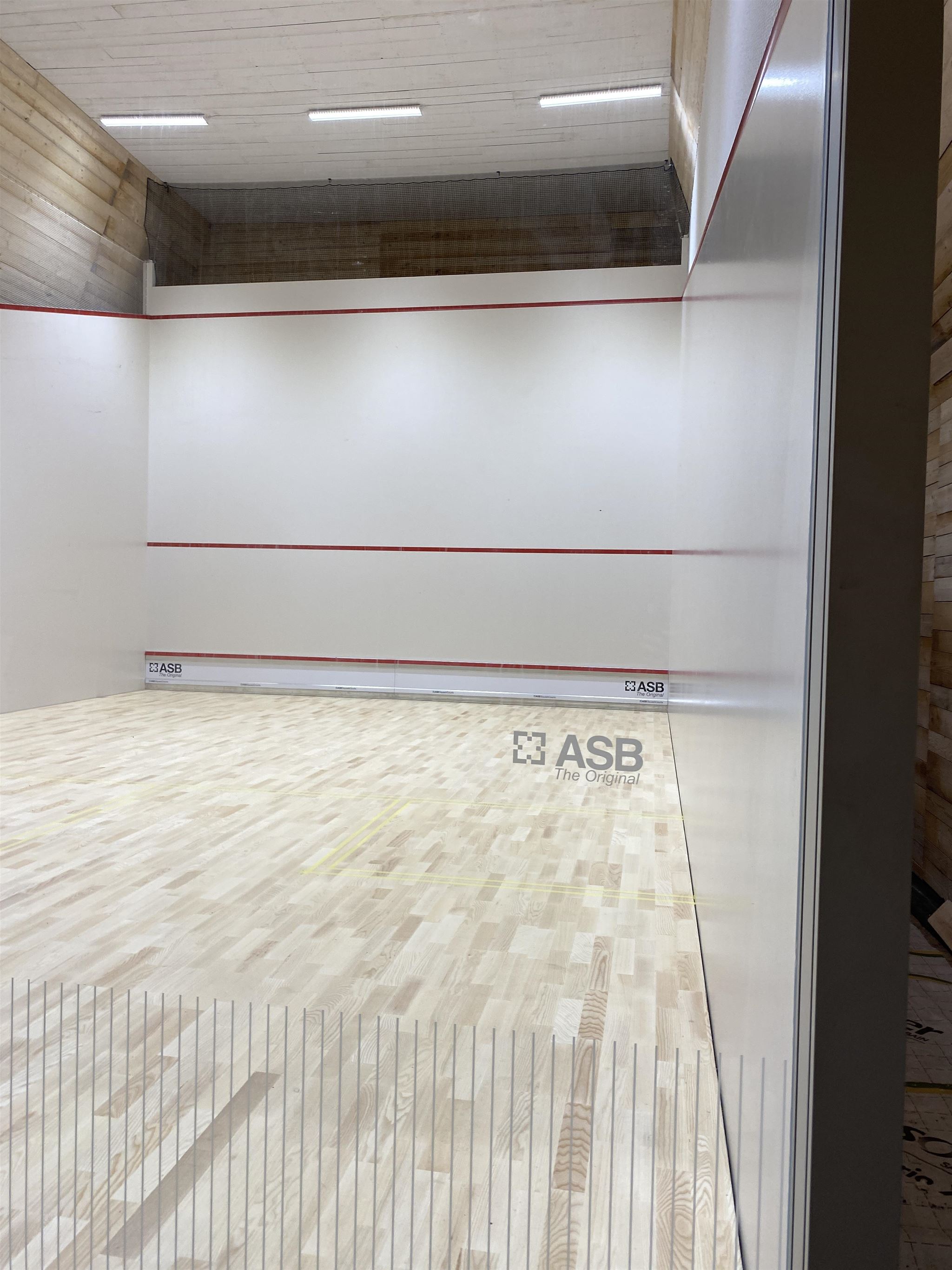 Ett modernt squashcourt med vita väggar, röda markeringslinjer och ljust trägolv.  Sokord: interiör, sport, squash, golvbeläggning, vägg, belysning · Sok aven: interior, indoor, lighting, lights
