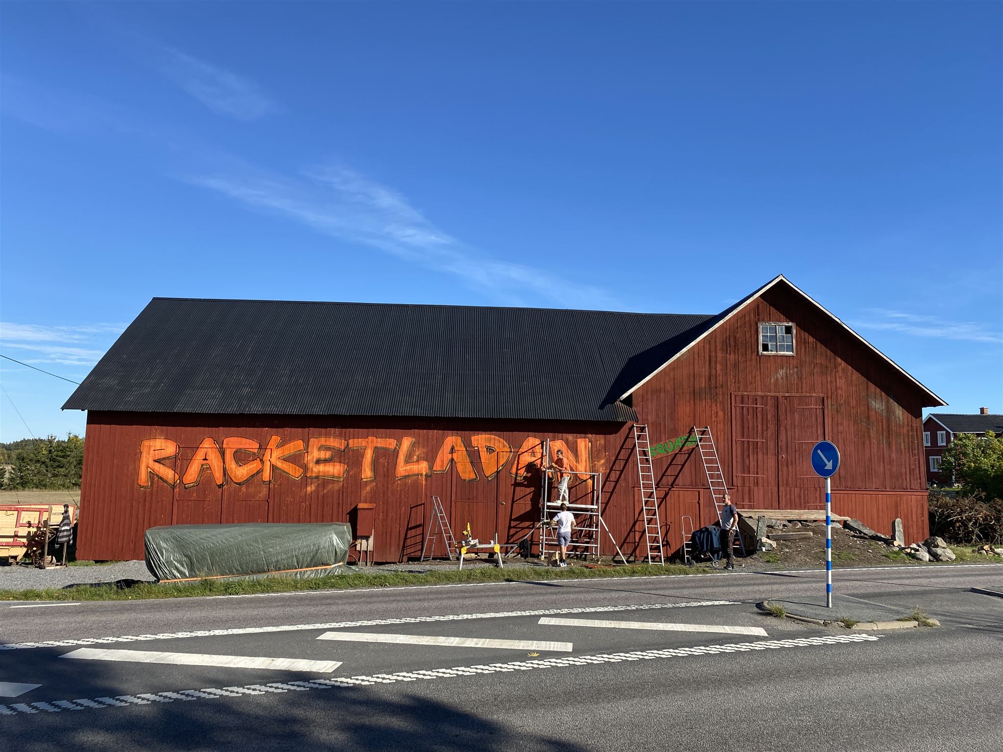 En röd lada med gul målad text och stegar lutade mot väggen.  Sokord: lada, röda hus, by, sommar, blå himmel · Sok aven: barn, red cottages, red houses, falu red, village, summer, blue sky